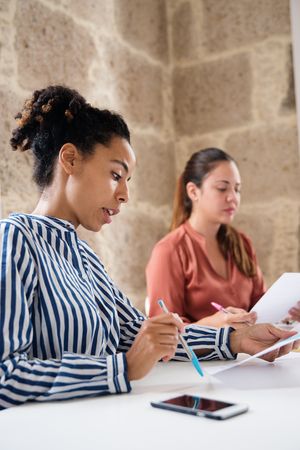 Two woman sitting in an office reading from papers that they are holding they both have a pen in their handsの写真素材