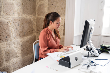 Woman with a long brown pony tail sitting in front of a computer typing as she sits at a deskの写真素材