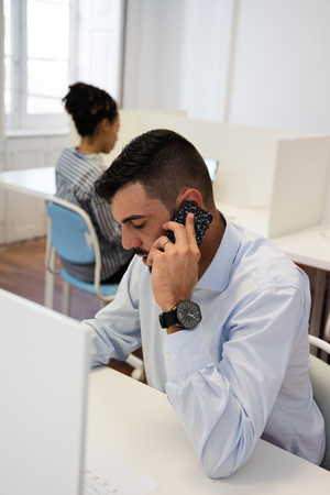 Man in a white shirt sits at a desk talking on a cell phone while looking downの写真素材