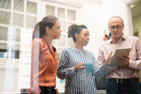 Three business team members standing in an office kitchen and discussing content displayed on a pc tabletの写真素材