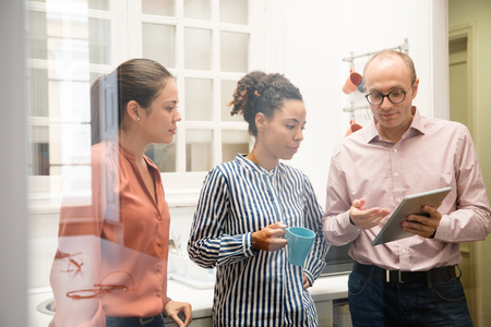 Three work colleagues standing in an office kitchen pointing and looking at a pc tabletの写真素材