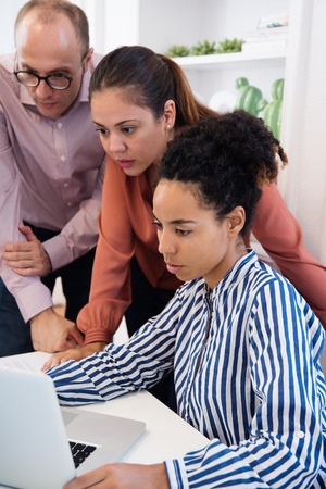 Group of office workers standing around a desk and looking at a computer with white shelves and green plantsの写真素材