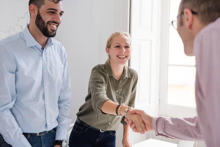 Man and woman in an office shake hands over a table and smile at each otherの写真素材