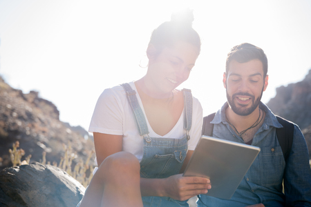 Smiling man sits on a rock next to a woman in the desert as they look at a pc tabletの写真素材