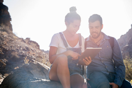 Man and woman sitting on a rock in a desert and they are smiling at a tablet pc screenの写真素材