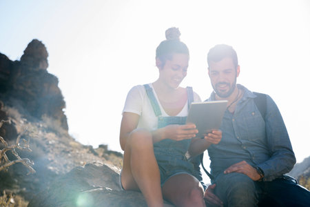 Couple sitting on a rock in the desert smiling as they look at a pc tablet screenの写真素材