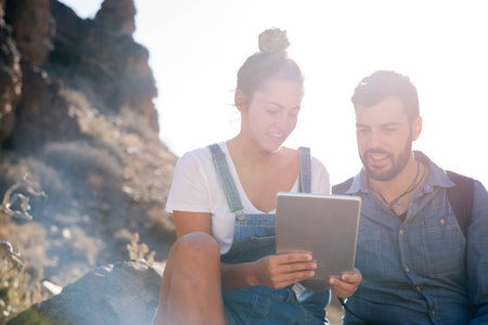 Smiling man sits next to a woman as she holds a pc tablet up, they are both sitting on a rock in a...