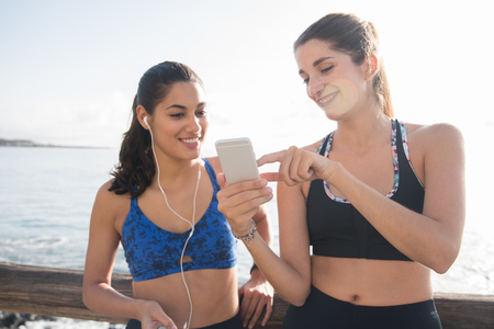 Two fitness females chatting and laughing while they are looking at a cell phone, they are standing at the beachの写真素材