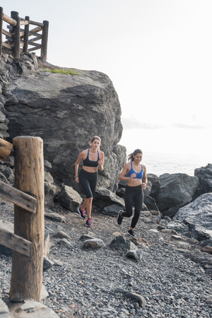 Women running on a dark rocky beach with large boulders and a wooden fence in the backgroundの写真素材