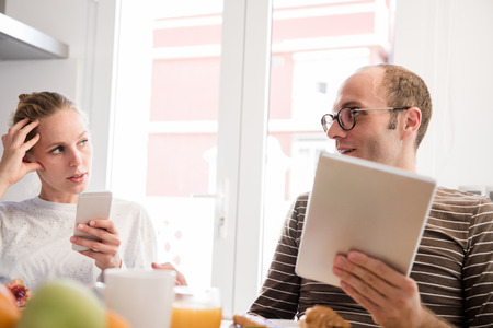 Couple sitting at a table talking and eating, she holds a cell phone and he is holding a pc tablet at breakfastの写真素材