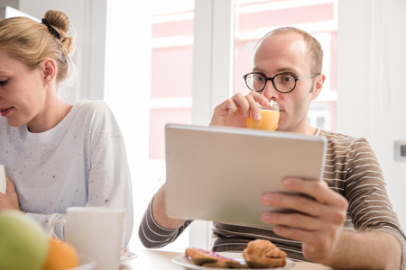 Man and woman sitting at a table, he is drinking a glass of juice and looking at a pc table at breakfastの写真素材