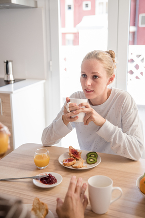 Woman sitting at a table having a breakfast and drinking from a mug as she look across at her companionの写真素材