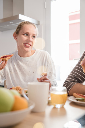 Woman sits at a table eating and smiles at her companion, they are in kitchen with white walls and large windowsの写真素材