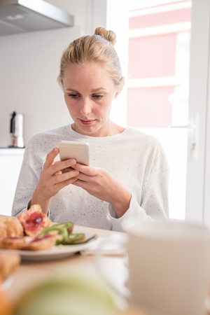 Woman sits at a table and holds a cell phone close to her face as she reads at breakfastの写真素材