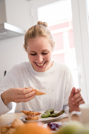 Woman eating a croissant and fruit, she is laughing and looking down at the food as she sits at breakfastの写真素材
