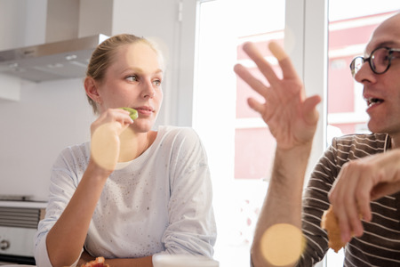 Woman eating kiwi fruit as she sits at a table in a kitchen as she listens to a man talking at breakfastの写真素材