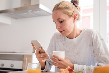 Close up of a blonde woman sitting at a table in a kitchen holding a white mug and texting on her cell phone at breakfastの写真素材