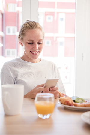 Woman at a breakfast table smiling and looking down at a cell phoneの写真素材
