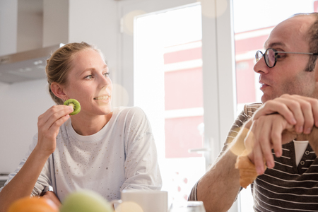 Couple sitting and talking as they look at each other at a kitchen table, she is eating fruit and he is eating a croissantの写真素材