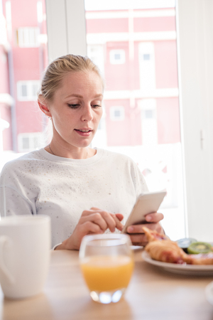 Woman sitting at a breakfast table looking down at her cell phone and pressing the screenの写真素材