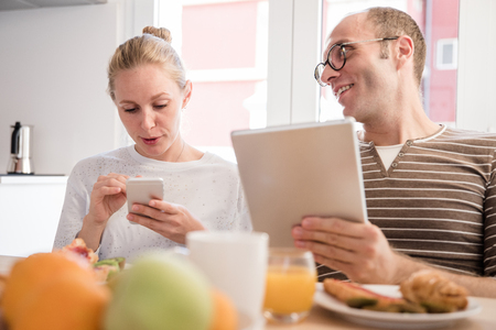 Couple laughing and talking as they sit at breakfast looking at their cellphones and a pc tabletの写真素材