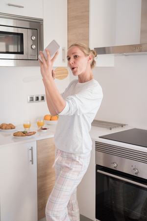 Blond woman standing in a kitchen as she takes a selfie with her cell phone and pretends to singの写真素材