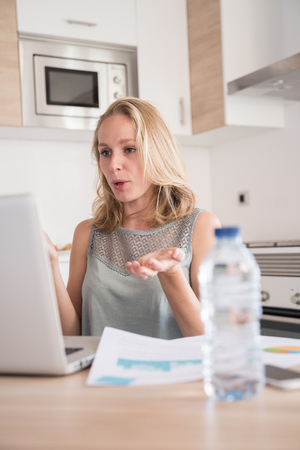 Woman sitting in a kitchen at a table as she gestures and talks to a laptop while working from homeの写真素材