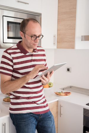 Man leaning against a kitchen counter as he holds a pc tablet as he textsの写真素材