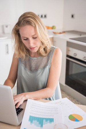 Woman sitting at a table in a kitchen typing on a laptop as she looks at documents next to her while working from homeの写真素材