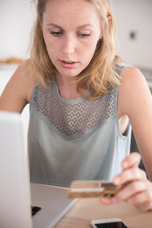 Woman looks at her credit cards and types on her pc laptop as she sits at a table while paying from homeの写真素材