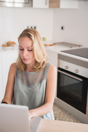 Blonde woman sitting at a table in a kitchen and typing on a laptop computer while working from homeの写真素材
