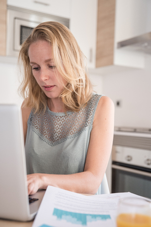Woman sitting at a table in a kitchen , she has her head down as she sits and types on a laptopの写真素材