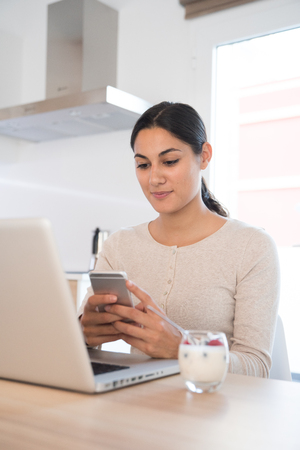 Woman looking at a cell phone as sits in front of a computer with a glass of yogurt and fruit in front of herの写真素材