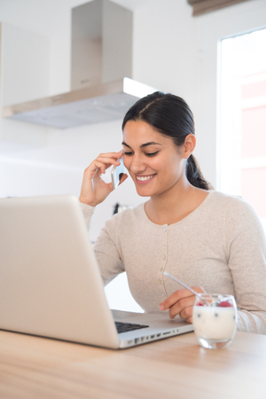 Woman talking on a cell phone and looking down and smiling as she sits at a table in front of a laptopの写真素材