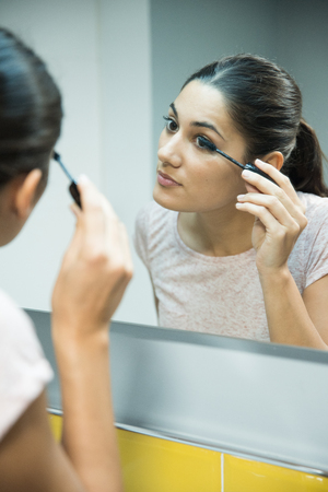 Woman leaning close to a mirror as she applies mascara to her eyelashesの写真素材