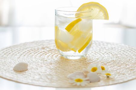 glass with a refreshing drink with ice and lemon on a white table with seashells and flowers