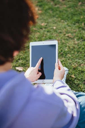 Girl with short hair is using a touch pad in the park. She is sitting in the grass.の写真素材