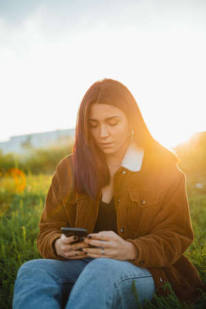 A teenager using her phone while sitting at a green meadow during the sunsetの写真素材