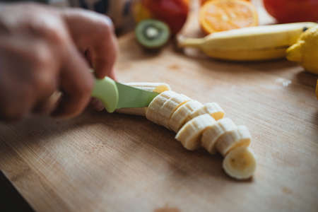 A woman cutting a banana with a green knife on a wooden table surrounded by other fruits.の写真素材