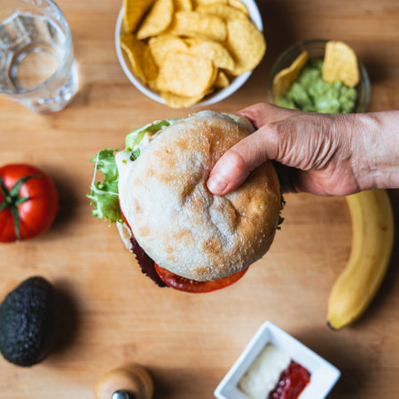 Fresh tasty burger with rustic bun surrounded by nachos with guacamole to dip in, a water glass, a tomato, an avocado, pepper, ketchup and mayonnaise and a banana.の写真素材