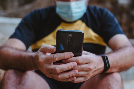 Close-up of a man wearing a mask looking at his phone while he is taking a break from running.の写真素材