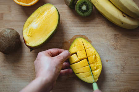 A woman cutting a mango with a green knife on a wooden table surrounded by other fruits (Top view).の写真素材