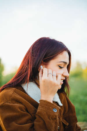 A teenager putting a white wireless ear-phone into her ear while sitting on the grass during sunsetの写真素材