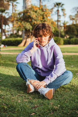 Sad girl wearing a purple hoodie talking on the phone in the park. She is sitting in the grass.の写真素材