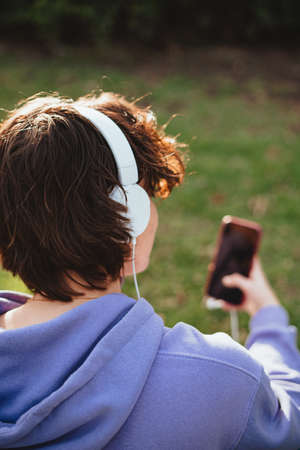 A teenager girl wearing white headphones to listen to music while sitting in the grass. She is using a smart phone.の写真素材