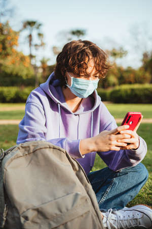 Student is using a mobile phone in the park. She is wearing a purple hoodie and a medical face mask.の写真素材