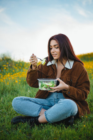 Teenager girl eating a salad while sitting on grass at a hill surrounded by yellow flowers. She is enjoying the calm.の写真素材
