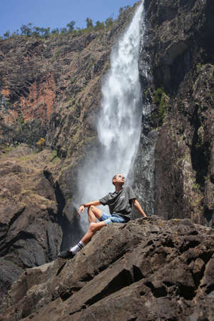 long hair man on a large rock under a huge waterfall in Australiaの写真素材