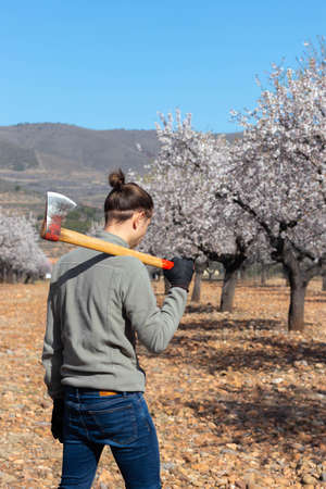 Young modern farmer with axe on his shoulder tree field in the background
