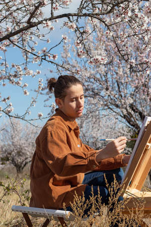 Young man practicing his hobby of painting in nature with beautiful scenery and landscape of flowers and fields aroundの写真素材